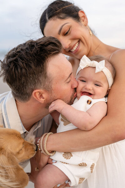 mother holding toddler while father kisses her and holds their family dog
