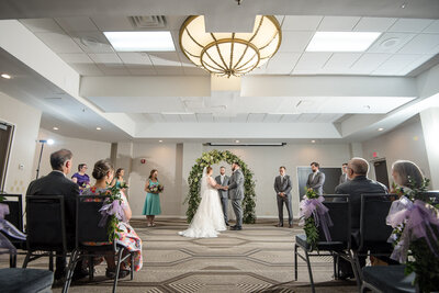 Indoor ballroom wedding ceremony at the Embassy Suites Hilton Charlotte where the bride and groom are holding hands and exchanging vows by Charlotte wedding photographers DeLong Photography