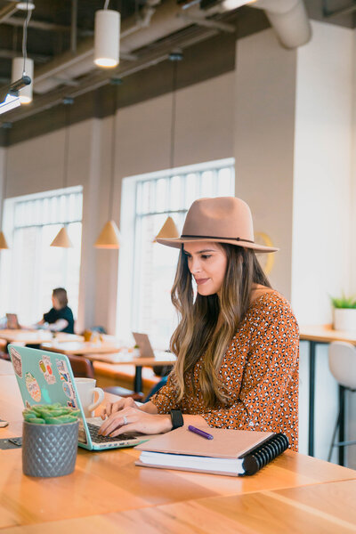 branding photographer captures young business owner sitting at a coffee shop typing on her laptop with a notebook to her side lightly smiling