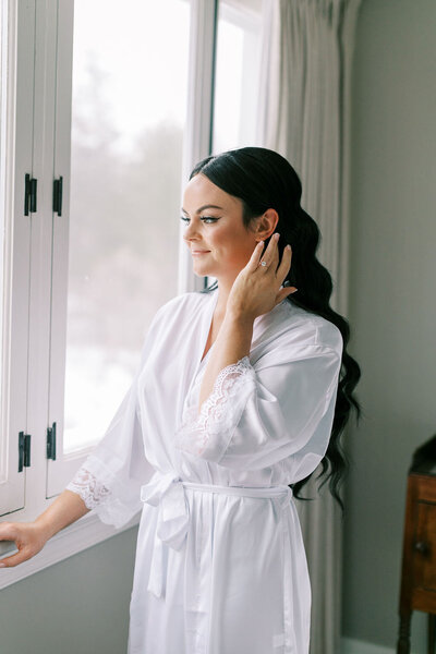 Close-up of a bride wearing an elegant lace wedding dress with floral details and sparkling drop earrings, softly touching her hair during a moment of reflection, captured by Twelfth Night Events in London, Ontario.