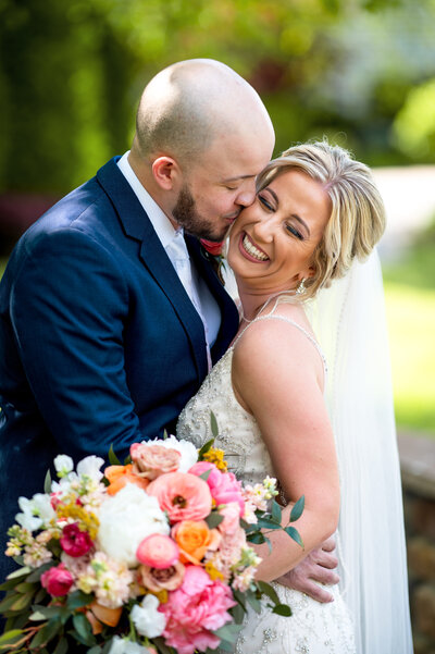 Groom kissing his bride on the cheek while she holds a colorful bouquet of flowers at their Riverton Country Club wedding