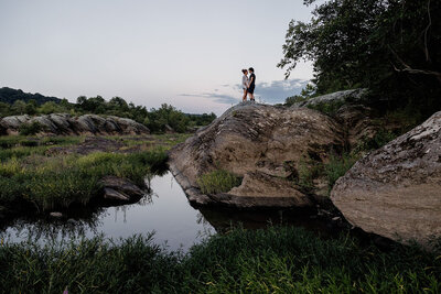 a couple stands on top of a rock surrounded by water. They are facing one another and holding hands