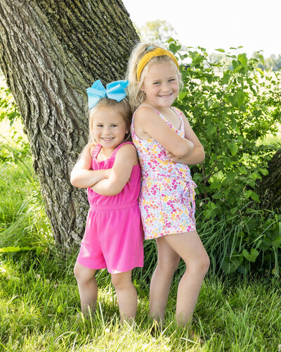 two young sisters standing back to back with arms crossed, looking at the camera smiling.  Younger sister has sandy blonde hair, wearing a pink jumper, and a blue bow in her hair.  Older sister has a white flower patterned one piece and a yellow headband
