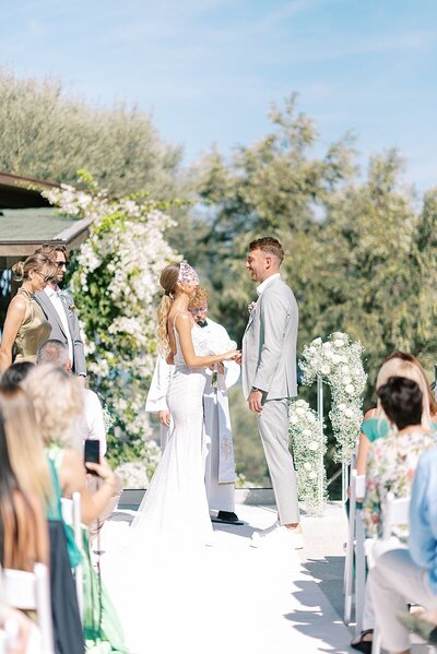 Newly married couple walk arm in arm under golden light, with the bride holding a bouquet and smiling at the groom during their destination wedding