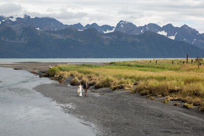 Bride and Groom walk hand in hand on a desolate black sand beach in on Resurrection Bay, Alaska, with giant snow-covered  mountains as a backdrop.