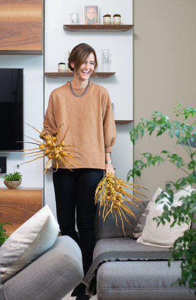 Sarah is stading in a living room with soft grey furniture. She is smiling and looking past the camera holding two intricate yellow spindly decor pieces.