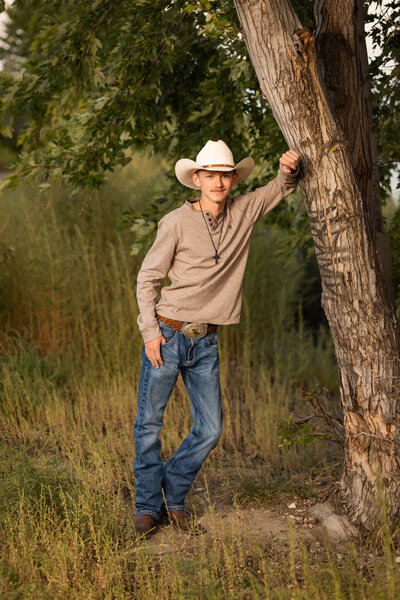high school senior boy in jeans and a tshirt wearing a cowboy hat leaning against a tree