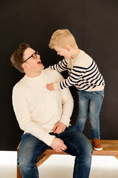 father sits on bench while toddler boy stands next to him on the bench, both looking at each other laughing at studio session in ann arbor michigan
