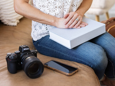 Close-up of photographer's hands resting on a linen photo album next to a Nikon camera and phone, showcasing heirloom products from a Kansas City photography session.