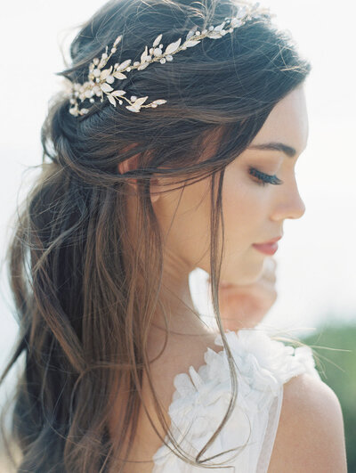 Kohnur ethereal bride with soft glam eye makeup and long loose waves styled with a gold leaf headpiece on a coastal cliffside in profile.