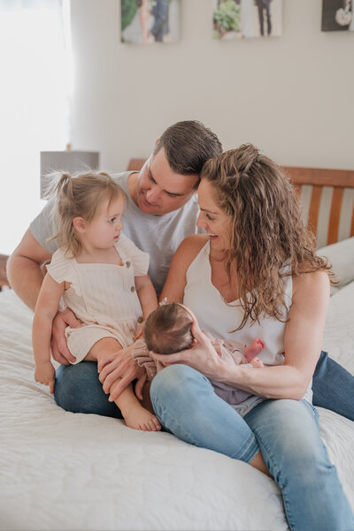 A family of four snuggles on a bed. Mom holds their newborn baby and she and dad are smiling at their toddler.