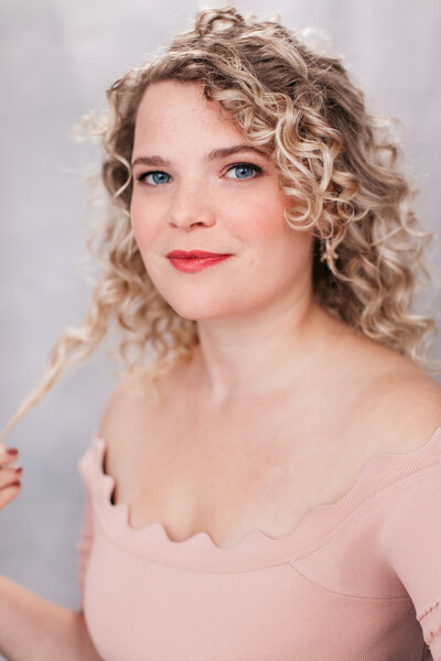 Castingfoto van zangeres Sylvia Boone. Speelse foto van vrouw met blonde krullen in roze top voor grijze achtergrond.