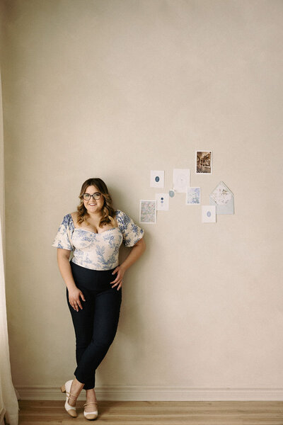 Wedding planner, Zelie & Co., leaning against a wall, smiling, with a wedding invitation flat lay on the wall
