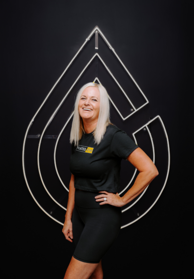 Headshot of Pilates instructor  standing in front of Pilates214 neon logo sign.