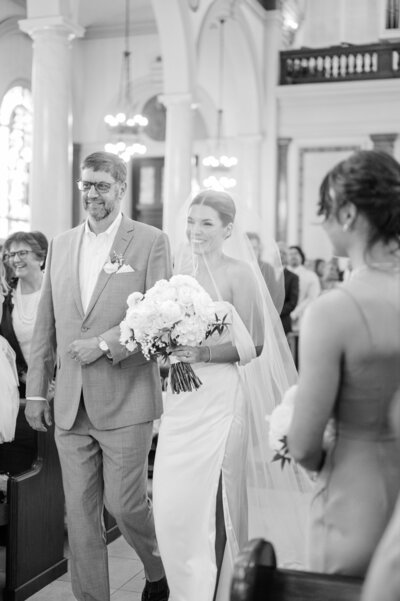 "Black-and-white image of a bride walking down the aisle at St. Mary's Catholic Church in Newport, Rhode Island, escorted by her father. The bride holds a bouquet of white flowers, smiling with joy as they approach the altar. Felisha, a New England-based destination wedding photographer, captures timeless moments at weddings in Newport, Rhode Island, New England, and worldwide locations.
