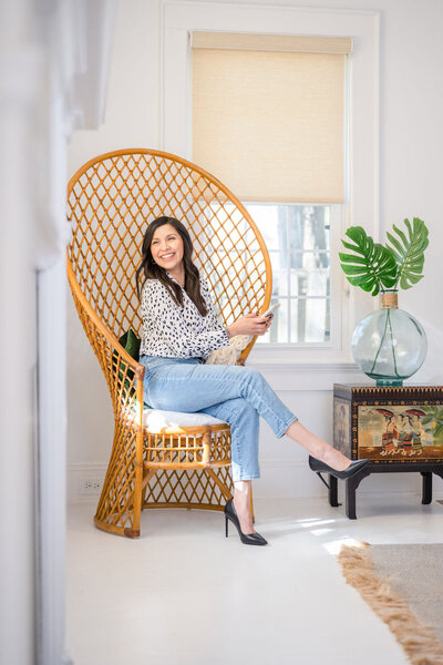woman sitting in a bamboo peacock chair wearing a black and white  blouse,  light jeans, black heels, and a smile as she crosses her legs and holds her phone in wilmington, north carolina