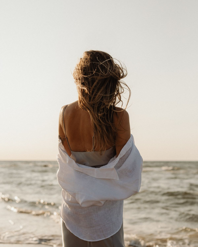 Woman wrapped in a white shirt standing at the edge of the ocean, her hair blowing in the wind during golden hour