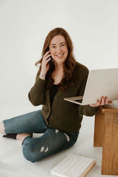 Virtually Robyn, a woman and virtual assistant, with long brown hair, wearing a green sweater, sits on the floor with a laptop, notebooks, and an iced drink, smiling at the camera while talking on the phone.