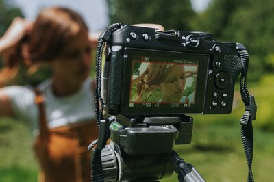 Camera sits on tripod shooting a picture of woman standing outdoors in a mustard-colored jumpsuit and white short-sleeve t-shirt.