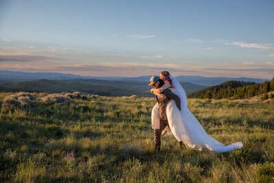 Groom gives his bride a piggyback ride in a meadow with the sunset turning the iconic Montana Big Sky into shades of pink and purple.