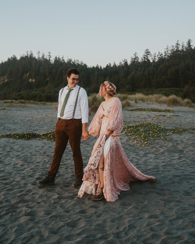 Big Sur elopement photography bride and groom walking down beach holding hands bride wearing rose pink lace wedding gown