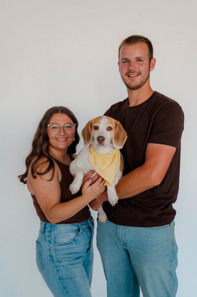 A couple stands in front of a white backdrop holding their dog and smiles at the camera.