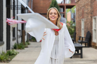 A joyful Loganville High School graduate in a white cap and gown tosses her cap into the air, smiling brightly in Downtown Monroe