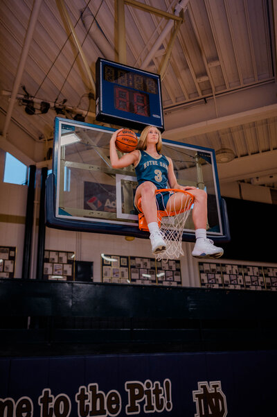 High school basketball player sits in basketball hoop with ball in hand.