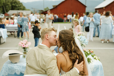 Wedding photography in Idaho Falls, Idaho by Adriana Montoya Photo. A groom kisses his bride while they are sitting at a table. Behind them are friends and family members at their reception. Behind them is a big red barn.