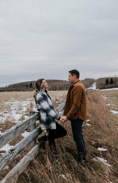 Woman leans against a fence in a Alberta foothills field while holding her fiance's hand and looking at each other lovingly