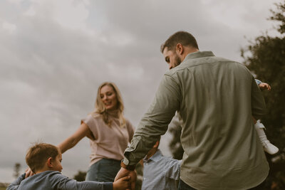 A young family holding hands running around in a circle with a stormy gray sky as their backdrop. 