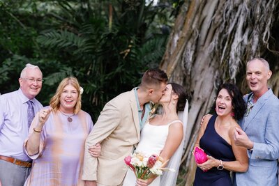 A newlywed couple kissing with their parents cheering next to them 