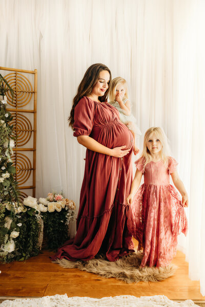 pregnant mom in rose maternity dress with 2 daughters - they are in a studio setting smiling at eachother