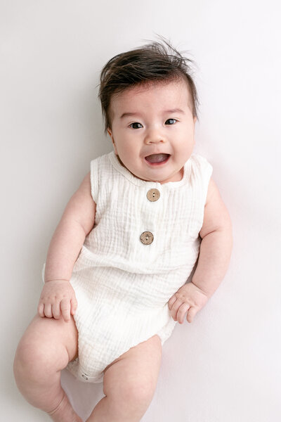 5 month old baby in white romper laying on white blanket smiling and looking right at camera.