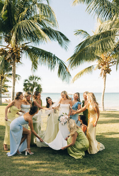 A group of bridesmaids in colorful dresses admire the bride and her gown on a beachside lawn under palm trees