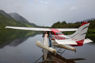 Bride and groom pose on the float of a red and white seaplane docked on a remote Alaskan lake, with mountains rising in the background. The groom wears a plaid shirt, while the bride’s wedding dress flows beside the aircraft, symbolizing an adventurous Alaska elopement.