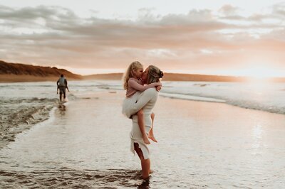 During  photo session a mom stands on a beach at sunset and hugs her young daughter and gives her a sweet kiss.