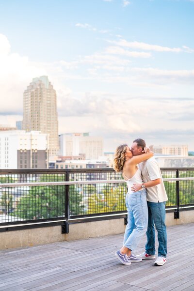 A man and a woman kissing with the cityscape of Raleigh, NC behind them.