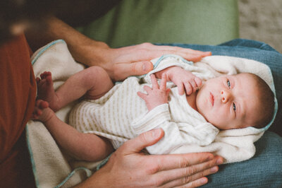 Newborn baby laying on  adult's lap with adults hands holding either side
