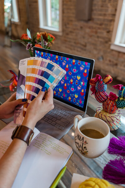 Digital artistry as the owner of our art workshops showcases a digitally crafted texture on the computer screen. Surrounding them are a Pantone printed with an array of colors, a cup of coffee, and artistic decorations