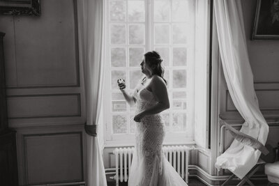 Black and white image of a bride in lace dress holding a wine glass while looking out a large vintage window.