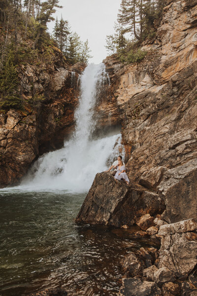 Couple sitting on a rock in front of a stunning waterfall in Montana.