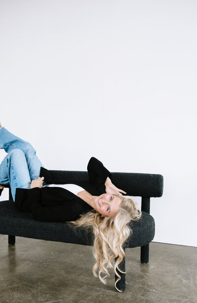 A joyful graduate in a white dress and blue stole poses playfully with a cap, one leg raised. A colorful bus and green trees are in the background.