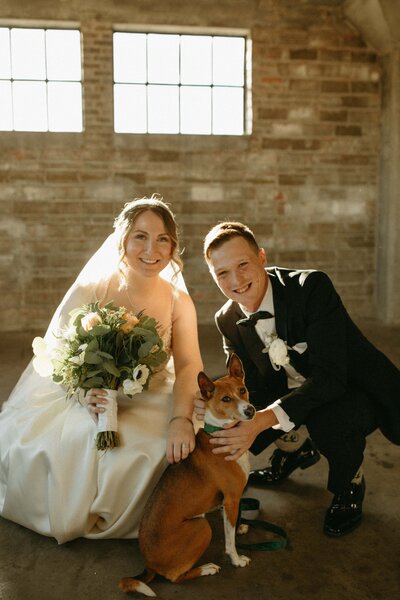 Bride and groom smiling with their dog in a sunlit industrial-style room, enjoying a stress-free wedding.