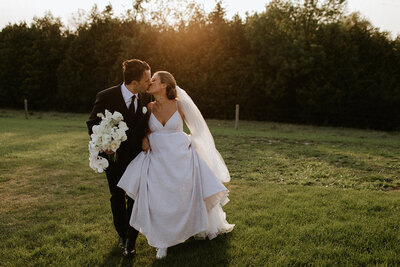 A bride and groom kiss while walking on grass, with the bride holding white flowers and the sun setting in the background—a perfect moment captured by a Toronto wedding photographer.