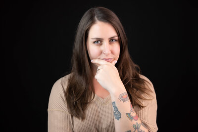 A family therapist with long brown hair and visible tattoos on her left forearm looks thoughtfully at the camera, resting her chin on her hand. Captured by a Jacksonville branding photographer, she wears a beige top against a plain black background.