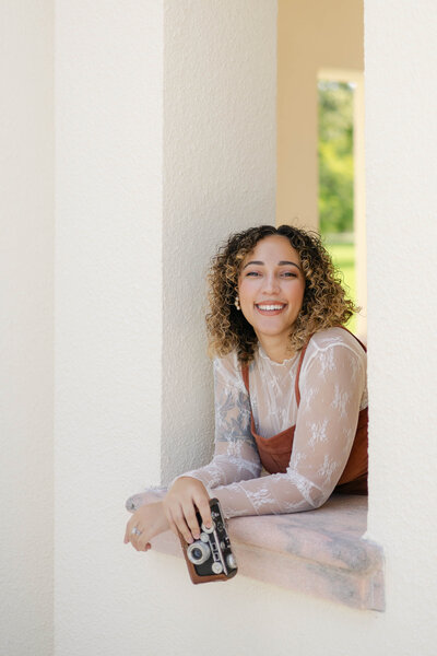 A photo of Zoe, the wedding photographer behind Popping Champaign Photography. She is smiling at the camera and holding a film camera. Zoe is a wedding photographer in Sarasota FL.