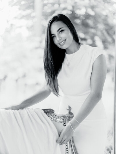 Black and white portrait of owner Linda Linares beside a vintage settee, smiling toward the camera.
