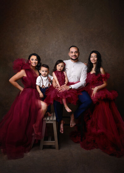 studio portrait of a family with 2 girls and a boy  with girls and mom wearing burgundy gowns and dad and son wearing white shirts with navy blue slacks