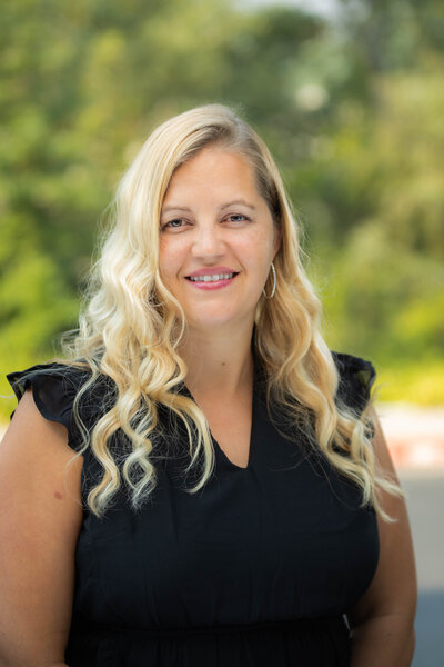 Woman poses for headshot with Sacramento headshot photographer in outdoor setting with greenery behind her. She is wearing a black shirt and wavy blond hair. She is smiling and looking directly at the camera.
